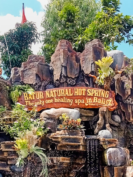 Batur Natural Hot Spring entrance sign with statue and lush greenery in Bali.