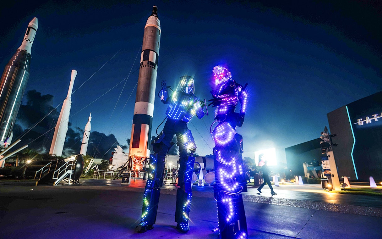 Illuminated performers at Kennedy Space Center after-hours event with rockets in the background.