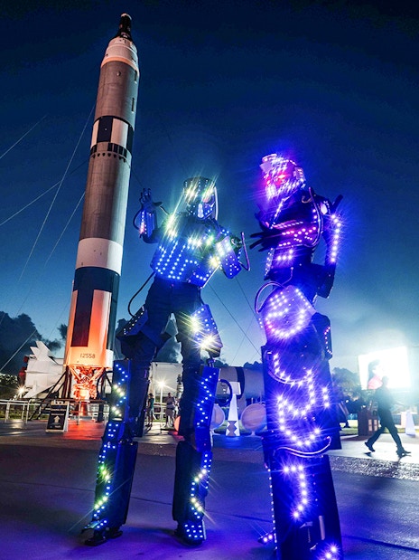 Illuminated performers at Kennedy Space Center after-hours event with rockets in the background.