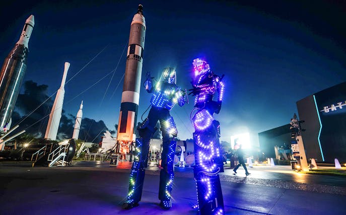 Illuminated performers at Kennedy Space Center after-hours event with rockets in the background.
