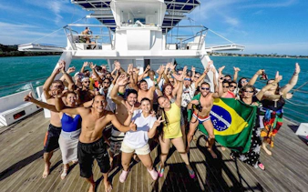 Tourists dancing on a boat during a DJ party, holding a Brazilian flag, ocean in the background.