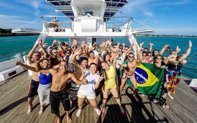 Tourists dancing on a boat during a DJ party, holding a Brazilian flag, ocean in the background.