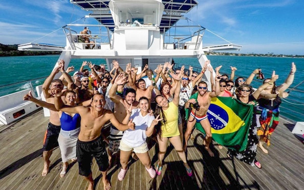 Tourists dancing on a boat during a DJ party, holding a Brazilian flag, ocean in the background.