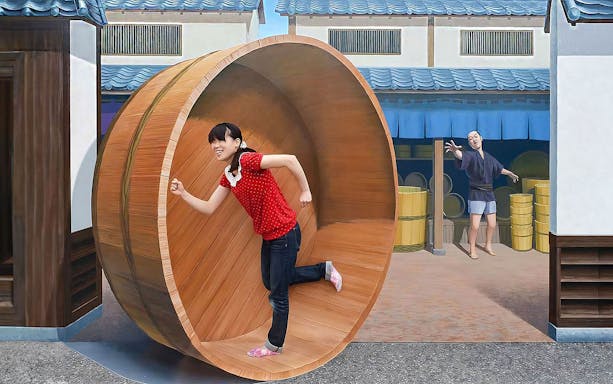 Person in a red shirt inside a large wooden wheel at Tokyo Trick Art Labyrinth Museum.