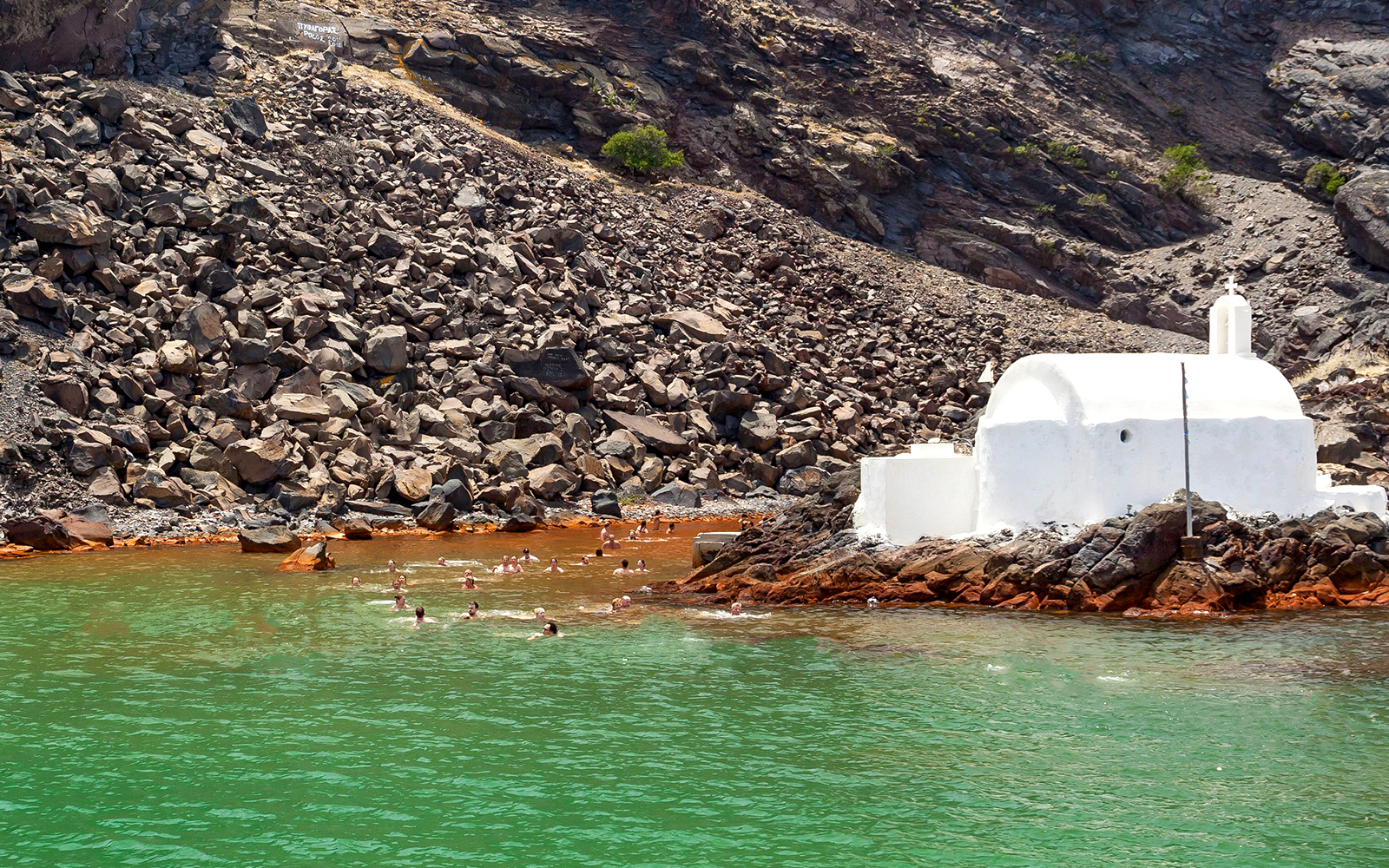 Swimmers near a white chapel on rocky shore, Santorini Private Sailing Catamaran Cruise.