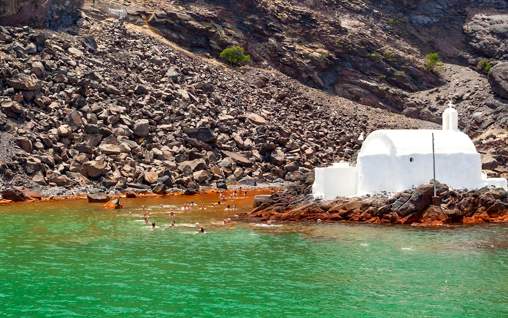 Swimmers near a white chapel on rocky shore, Santorini Private Sailing Catamaran Cruise.