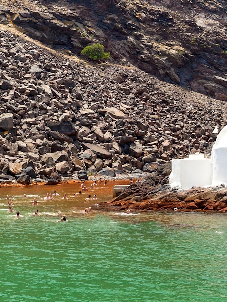 Swimmers near a white chapel on rocky shore, Santorini Private Sailing Catamaran Cruise.