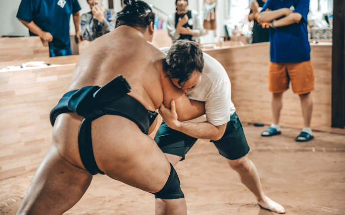 Participant in white t-shirt grappling with sumo wrestler in a ring.
