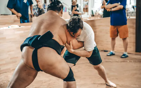 Participant in white t-shirt grappling with sumo wrestler in a ring.