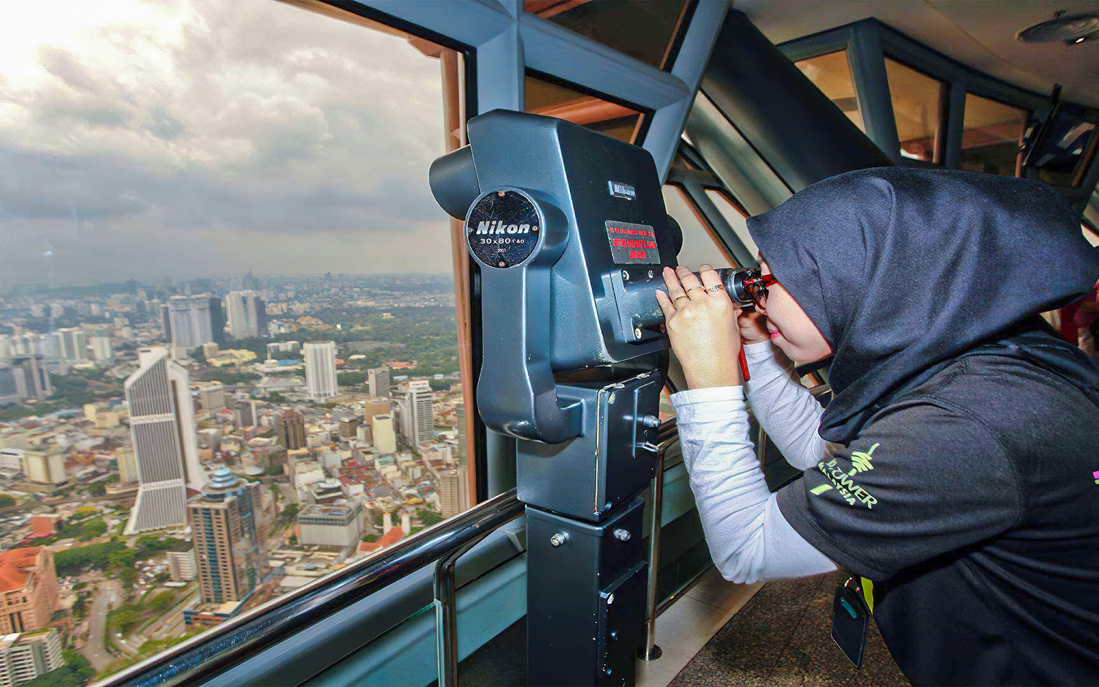 Visitor using binoculars at KL Tower observation deck overlooking Kuala Lumpur skyline.