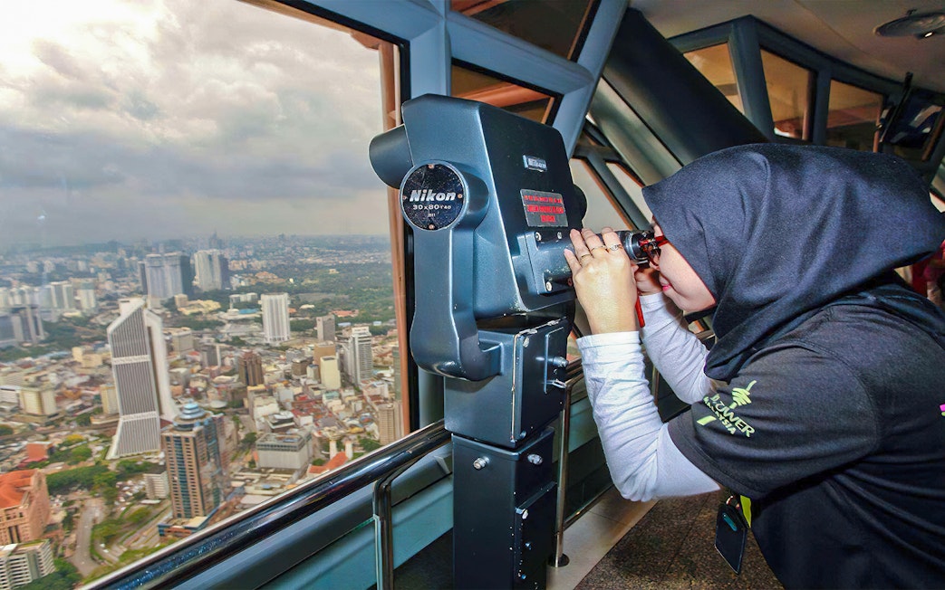 Visitor using binoculars at KL Tower observation deck overlooking Kuala Lumpur skyline.