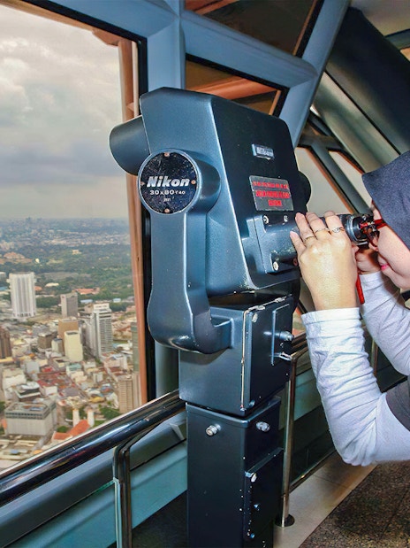 Visitor using binoculars at KL Tower observation deck overlooking Kuala Lumpur skyline.