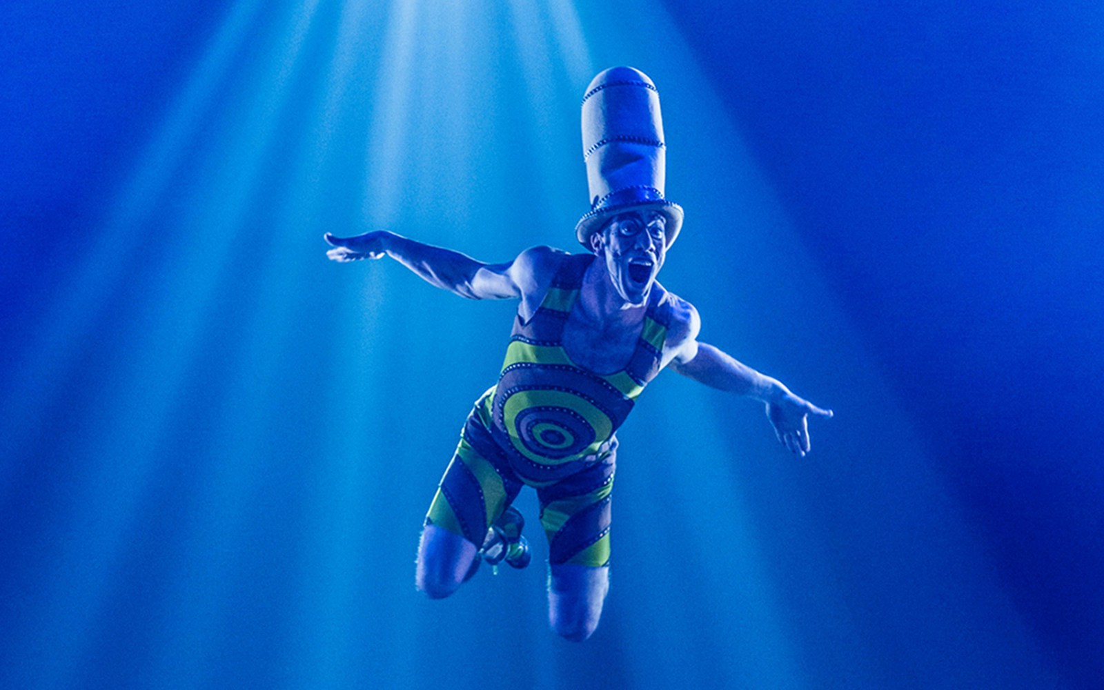Performer in Cirque du Soleil: Kurios, wearing a striped costume and tall hat, mid-air under blue lights.