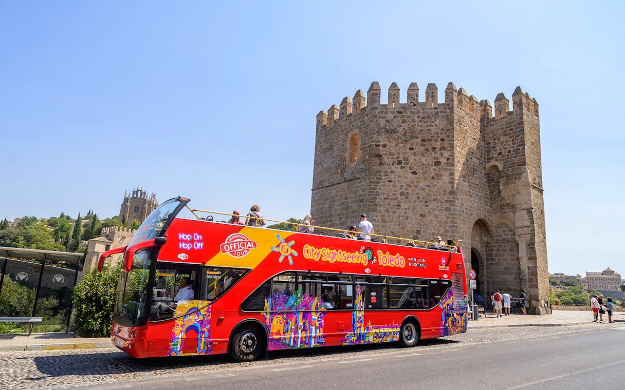 Red double-decker bus on Toledo Hop-On Hop-Off Tour near historic stone gate.
