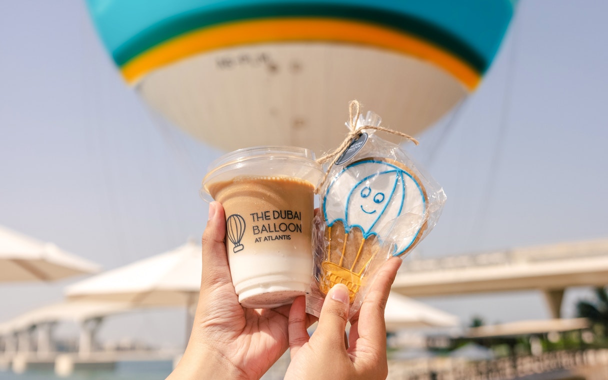 Hands holding a drink and cookie with The Dubai Balloon at Atlantis in the background.
