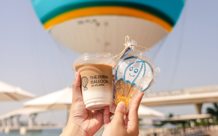 Hands holding a drink and cookie with The Dubai Balloon at Atlantis in the background.