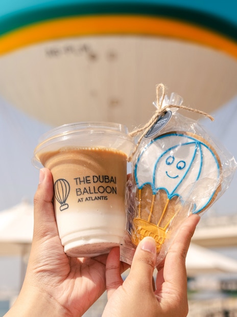 Hands holding a drink and cookie with The Dubai Balloon at Atlantis in the background.