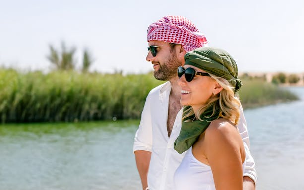 Tourists by a river wearing Ghutra headscarves in a sunny landscape.