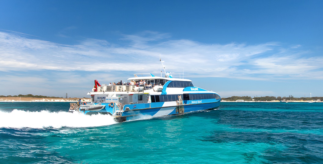 Eine Gruppe von Touristen genießt einen sonnigen Tag auf der Rottnest Island Guided Bus Tour, mit Blick auf die Küste und die Fähre, die am Dock in Perth oder Fremantle auf die Rückfahrt wartet.