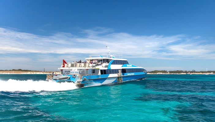 Group of tourists enjoying a sunny day on the Rottnest Island Guided Bus Tour, with a scenic view of the coastline and the return ferry waiting at the dock in Perth or Fremantle