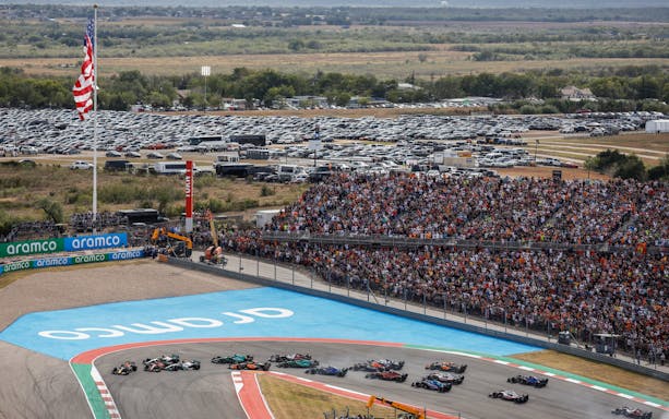 Formula 1 cars racing at the USA Grand Prix with a large crowd and American flag in view.