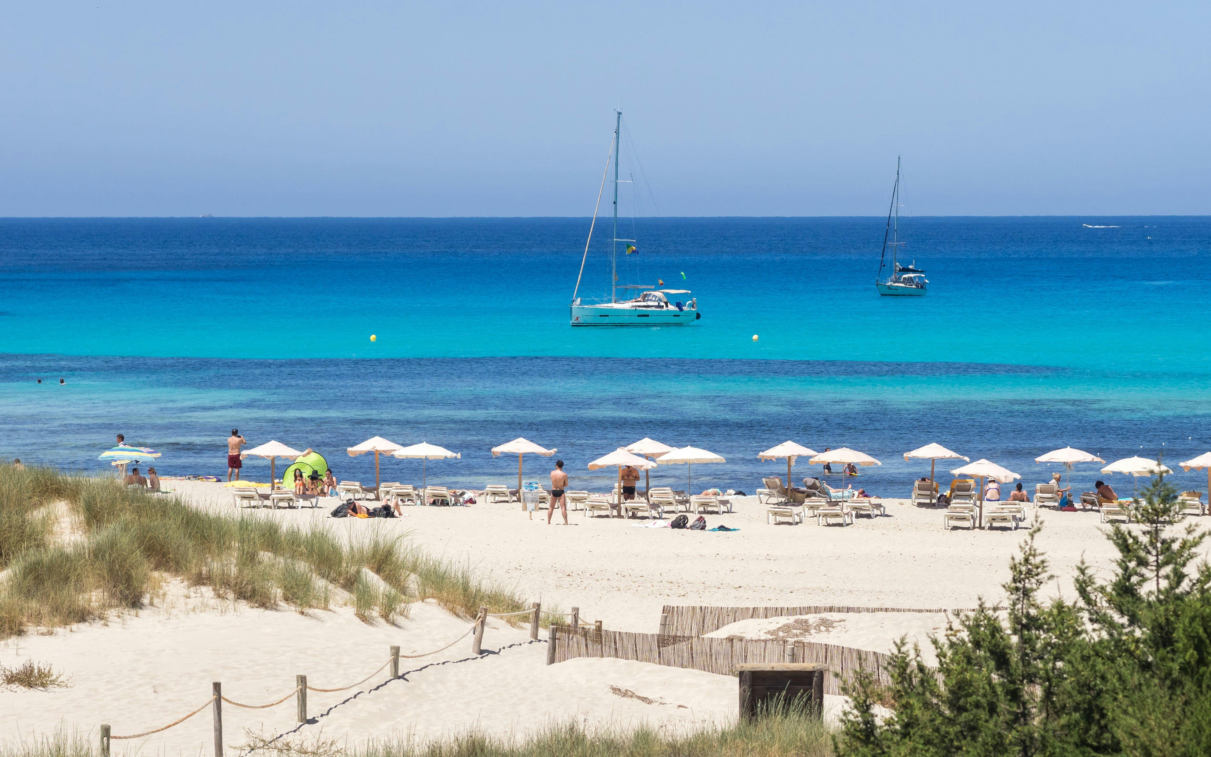 Sandy beach with sun loungers and umbrellas at Cala Saona, Formentera, with sailboats on turquoise water.
