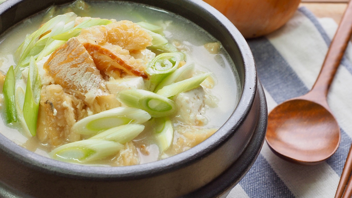 Korean fish soup with green onions in a black bowl on a striped cloth.