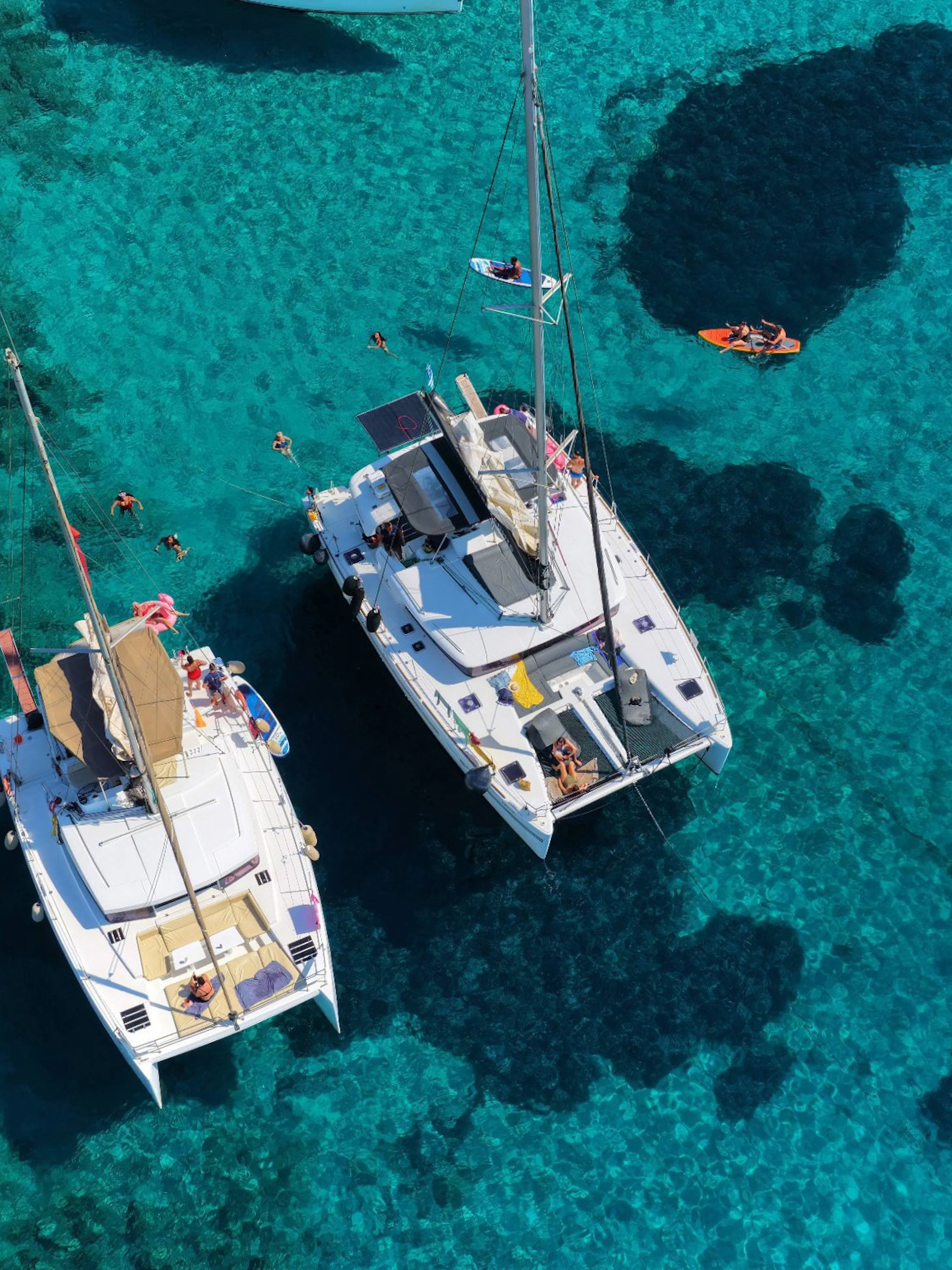 Catamarans anchored near Dia Island, Crete, with people swimming and paddleboarding in clear waters.
