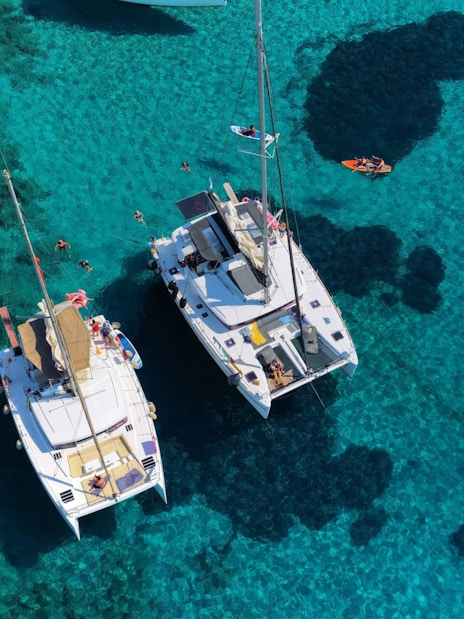 Catamarans anchored near Dia Island, Crete, with people swimming and paddleboarding in clear waters.