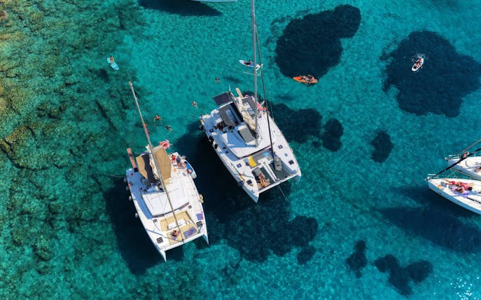 Catamarans anchored near Dia Island, Crete, with people swimming and paddleboarding in clear waters.