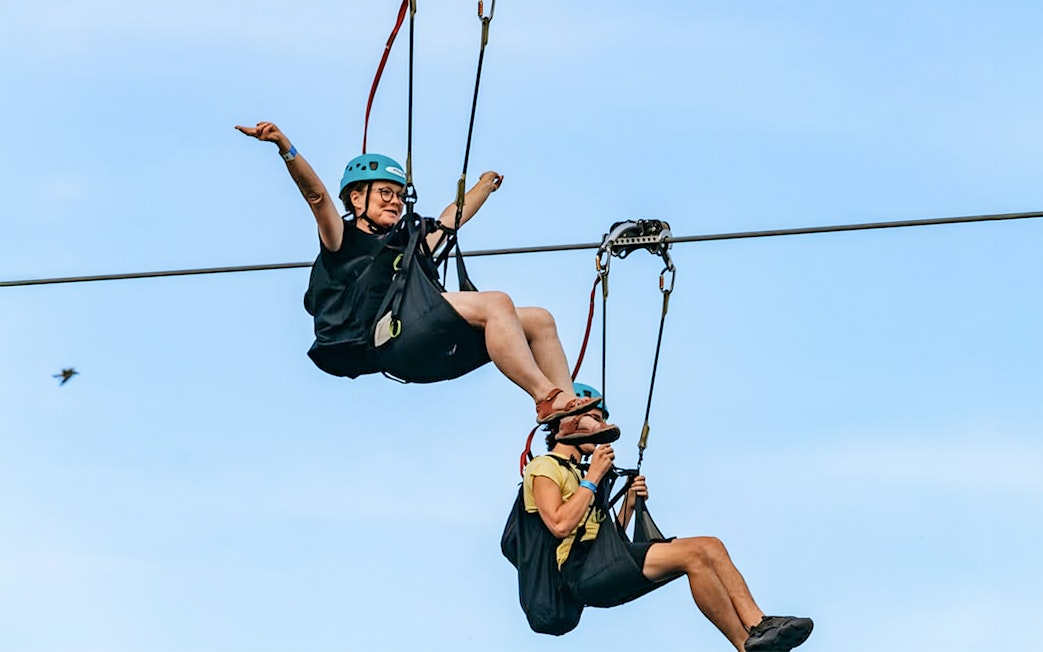 Two people ziplining with helmets and harnesses against a clear sky, part of the Zipline To The Falls experience.