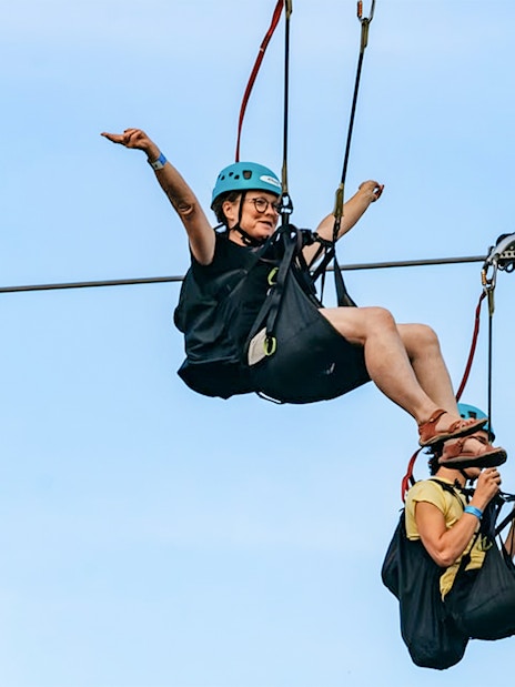 Two people ziplining with helmets and harnesses against a clear sky, part of the Zipline To The Falls experience.