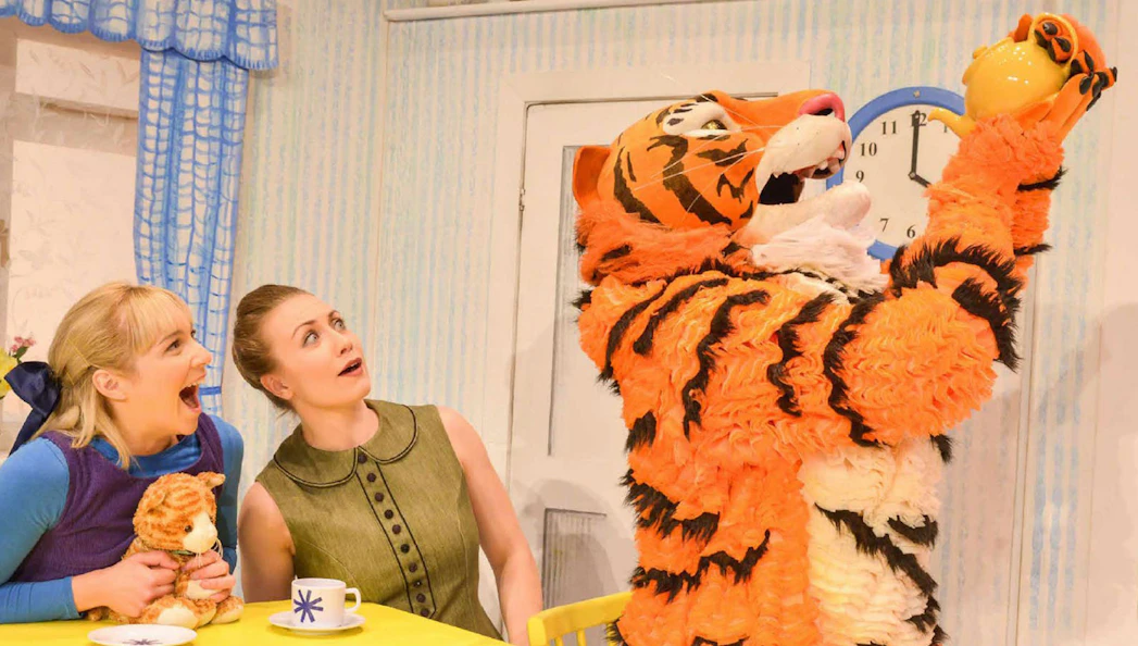 Tiger character holding teapot with two surprised women at a table, from "The Tiger Who Came to Tea" show.