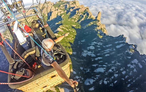Tourists in a hot air balloon over mountainous landscape with clouds.