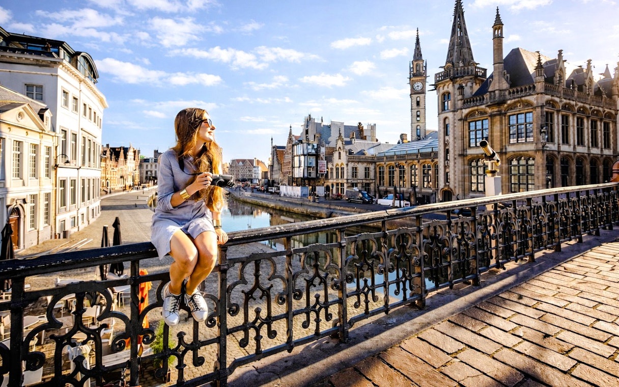 Person sitting on Sint-Michielsbrug in Ghent, holding a camera with historic buildings in the background.