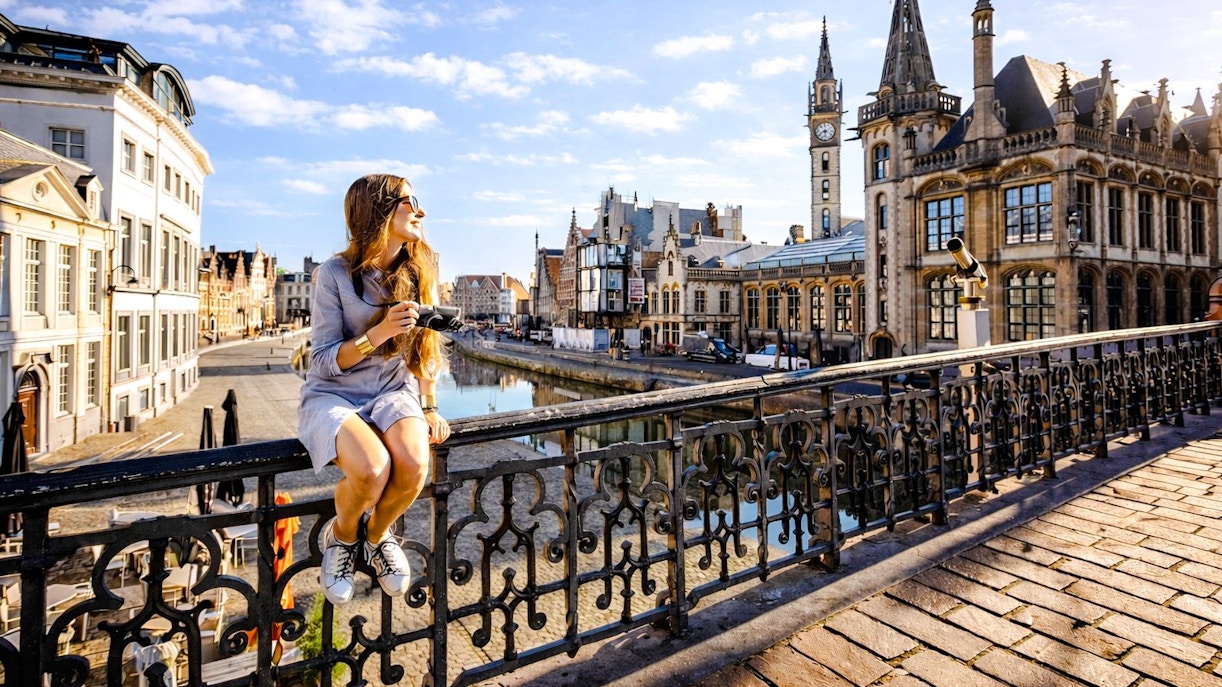 Person sitting on Sint-Michielsbrug in Ghent, holding a camera with historic buildings in the background.