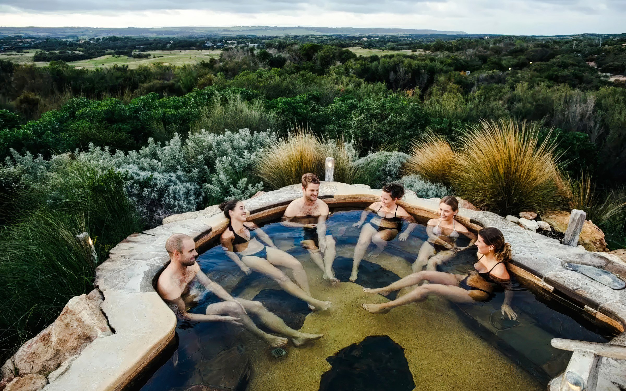 Group relaxing in a hot spring pool at Peninsula Hot Springs, surrounded by lush greenery.