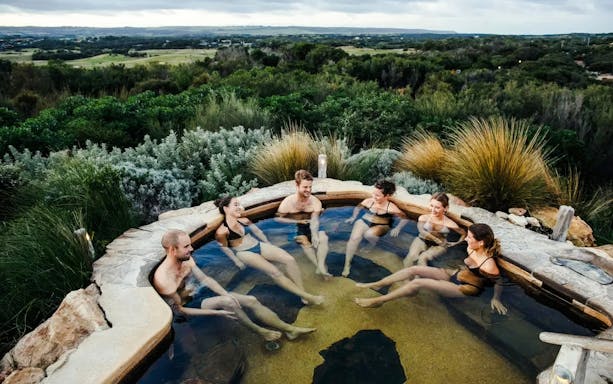Group relaxing in a hot spring pool at Peninsula Hot Springs, surrounded by lush greenery.