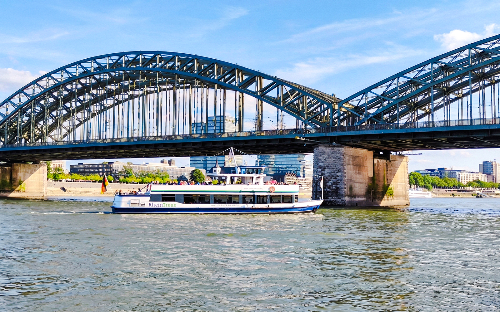 Cruise boat on the Rhine River passing under Hohenzollern Bridge, Cologne city skyline in background.