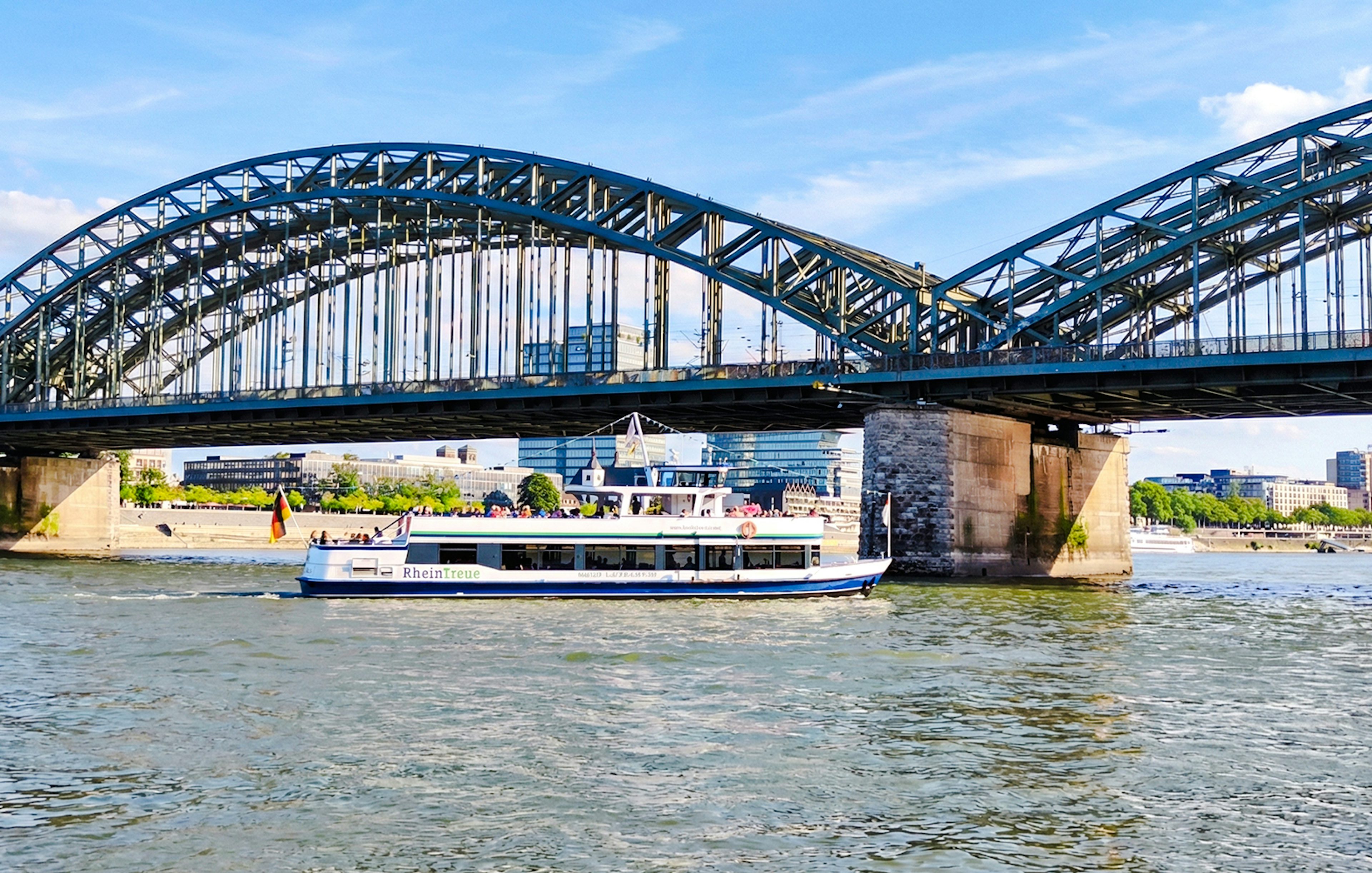 Cruise boat on the Rhine River passing under Hohenzollern Bridge, Cologne city skyline in background.