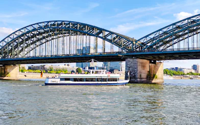Cruise boat on the Rhine River passing under Hohenzollern Bridge, Cologne city skyline in background.