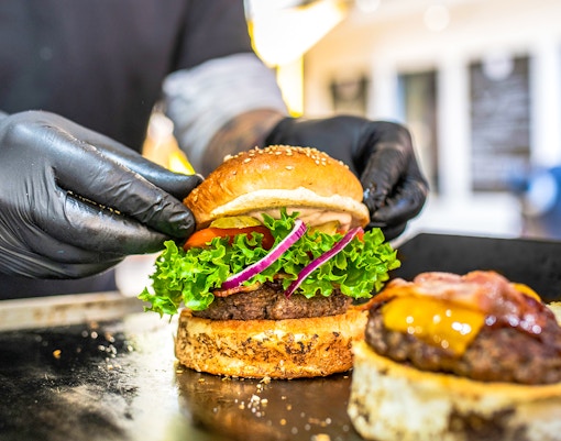 Chef assembling gourmet burger with lettuce, tomato, and onion on a grill.