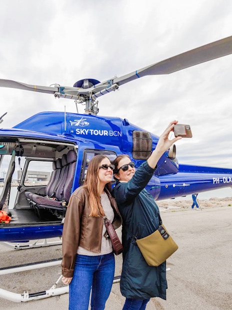 Tourists taking a selfie near a blue helicopter in Barcelona.