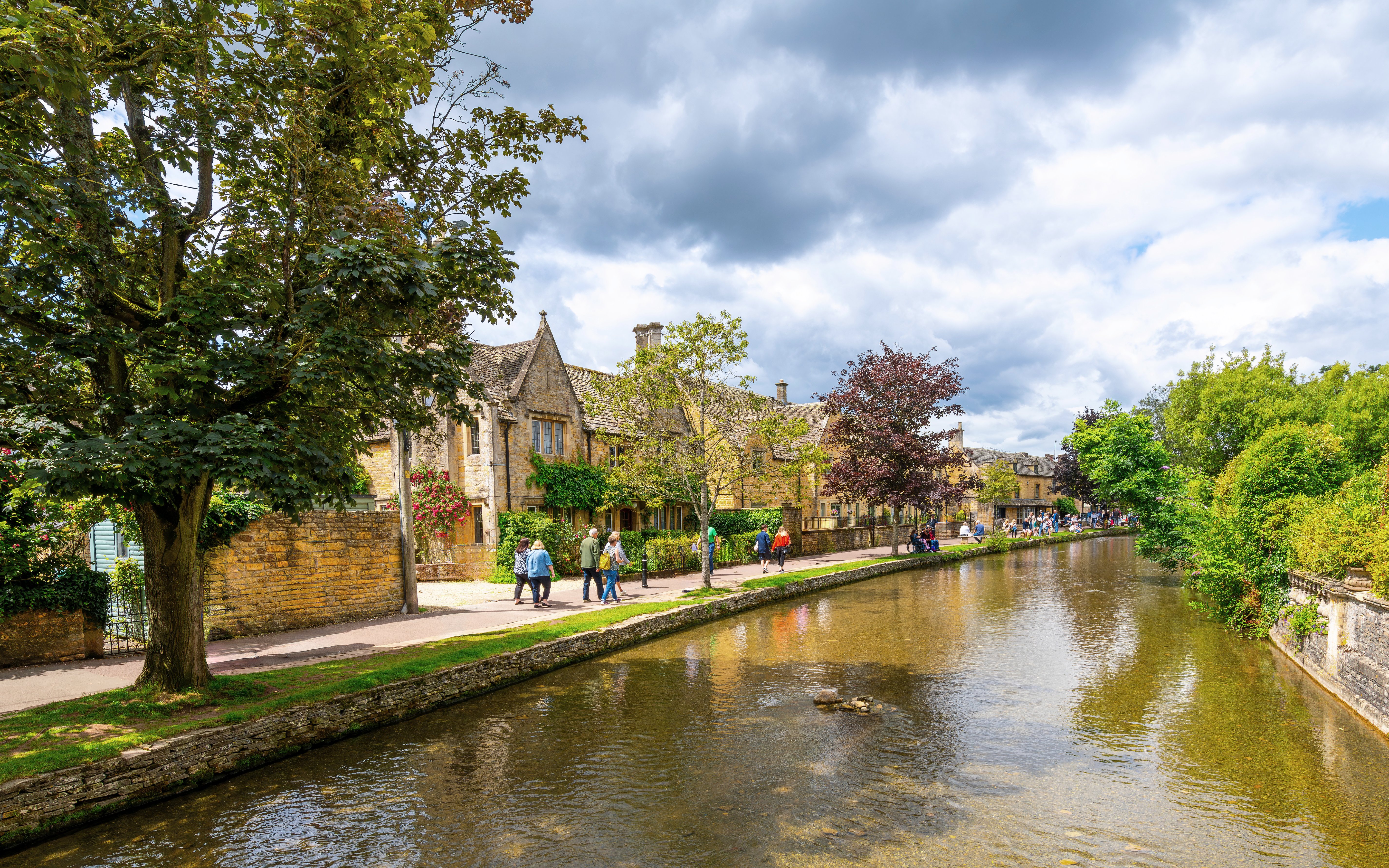 Riverside path with people walking in Bourton-on-the-Water, Cotswolds.
