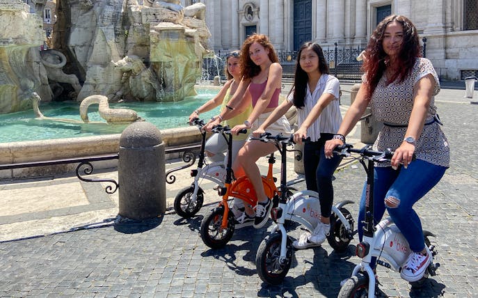 Visitors on e-bikes near a historic fountain in Rome, Italy, for Circus Maximus virtual reality tour.