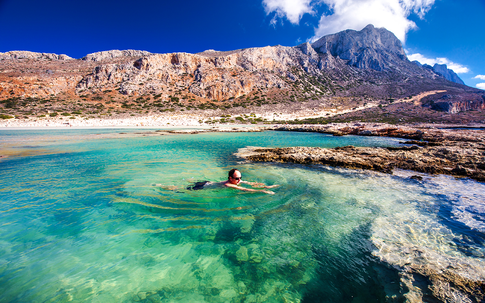 Man swimming in clear waters of Balos Lagoon, Crete, with rocky landscape in background.