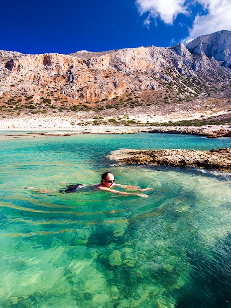 Man swimming in clear waters of Balos Lagoon, Crete, with rocky landscape in background.