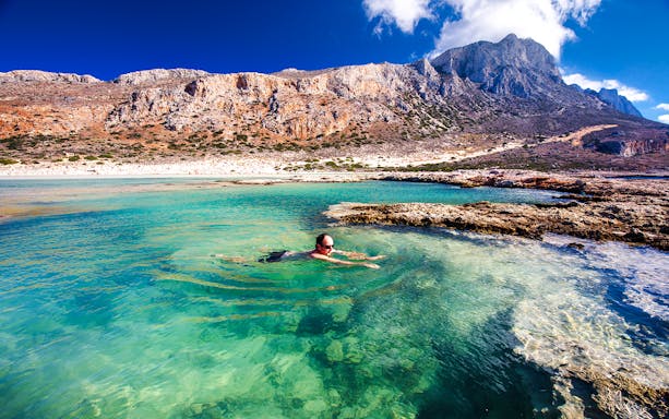 Man swimming in clear waters of Balos Lagoon, Crete, with rocky landscape in background.