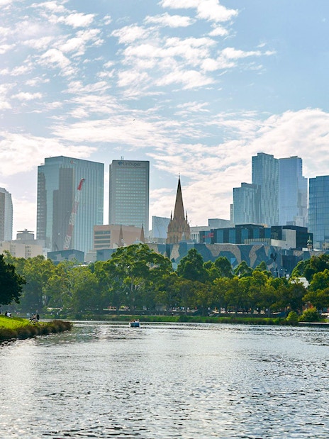 Melbourne skyline viewed from Yarra River, featuring cityscape and riverbank greenery.