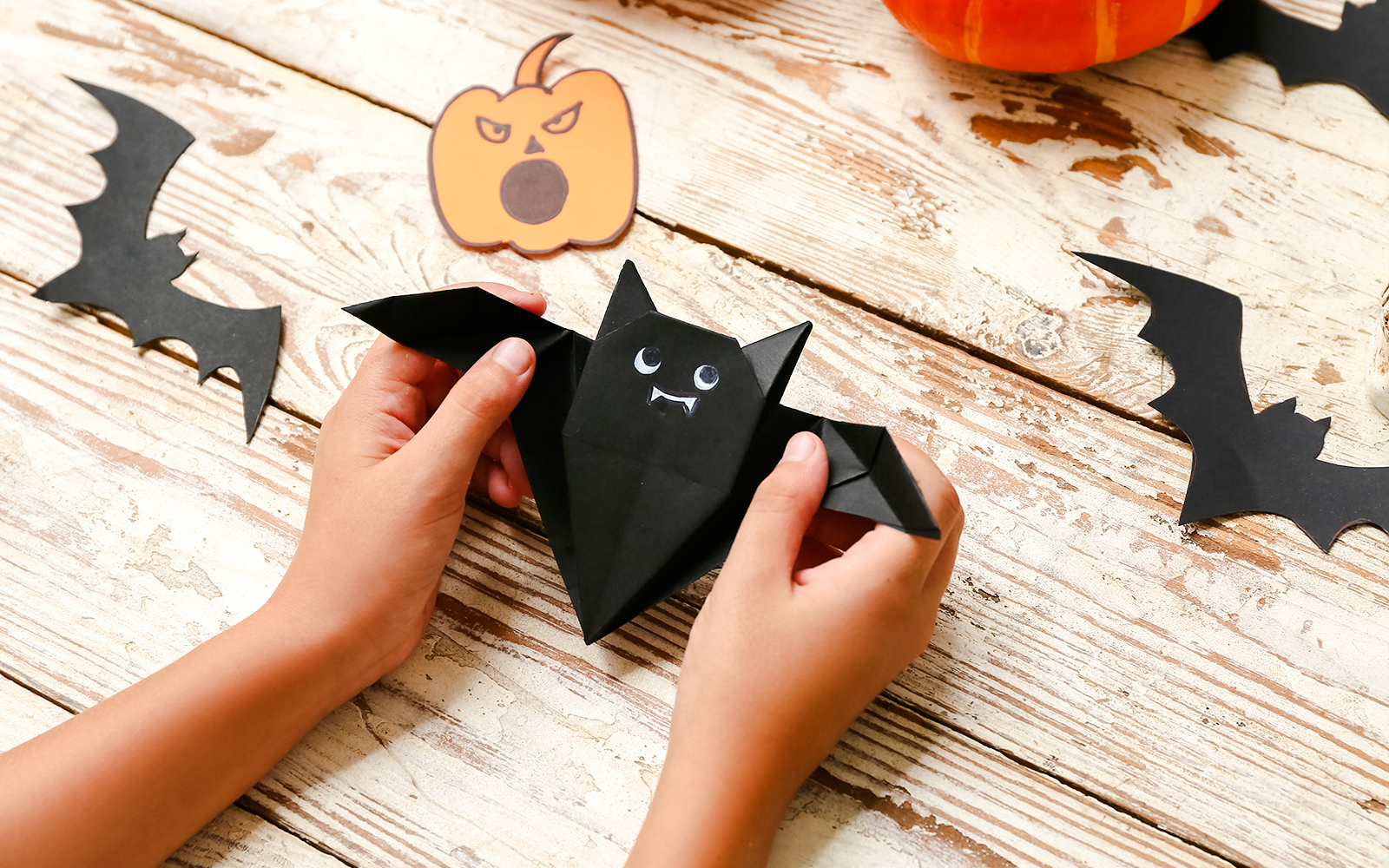 Child crafting origami bat with Halloween decorations on wooden table.
